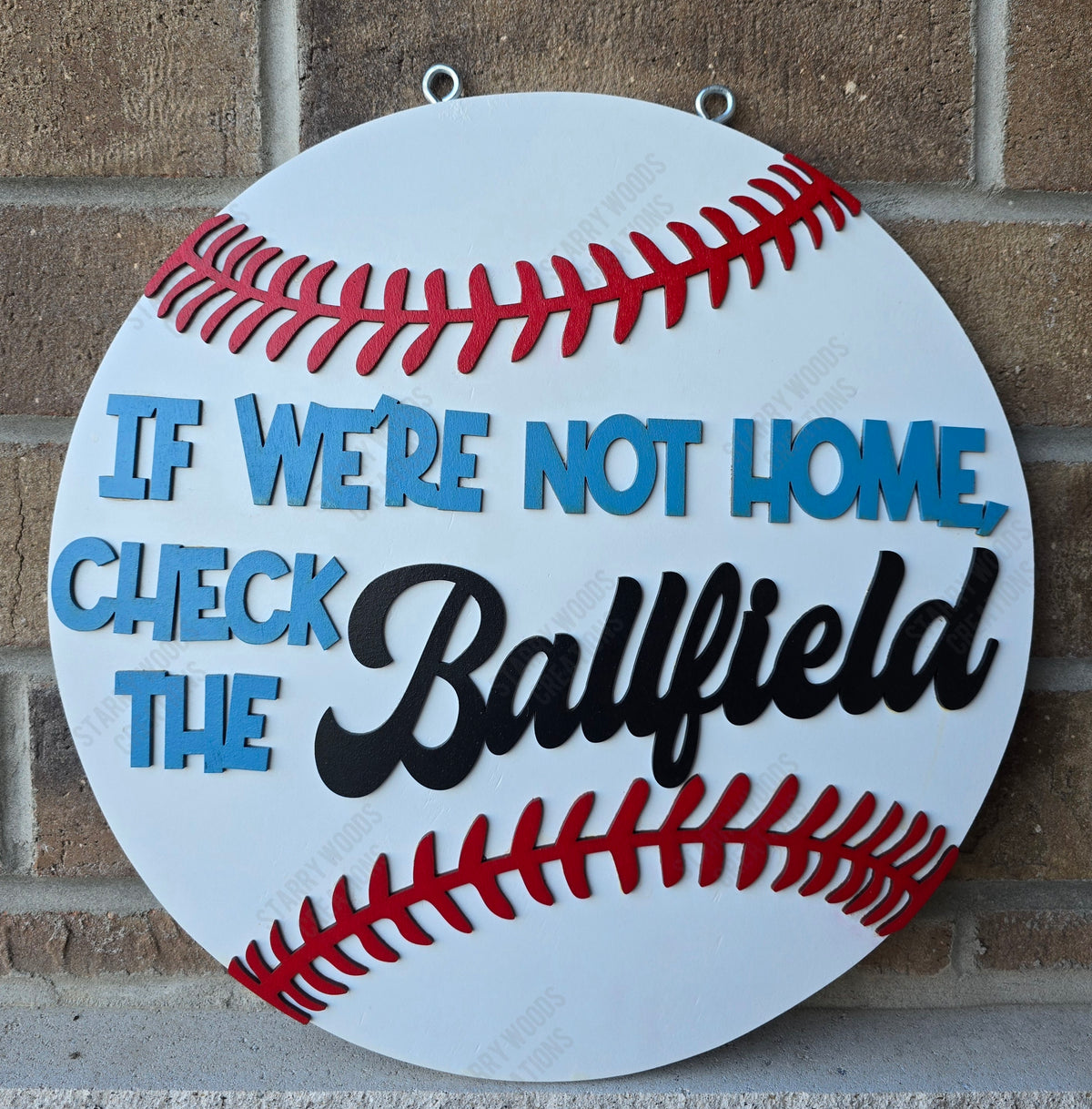 Wooden sign shaped like a baseball with text 'If we're not home check the ballfield' on a brick wall background. Handmade in Quinte West and ready to hang.