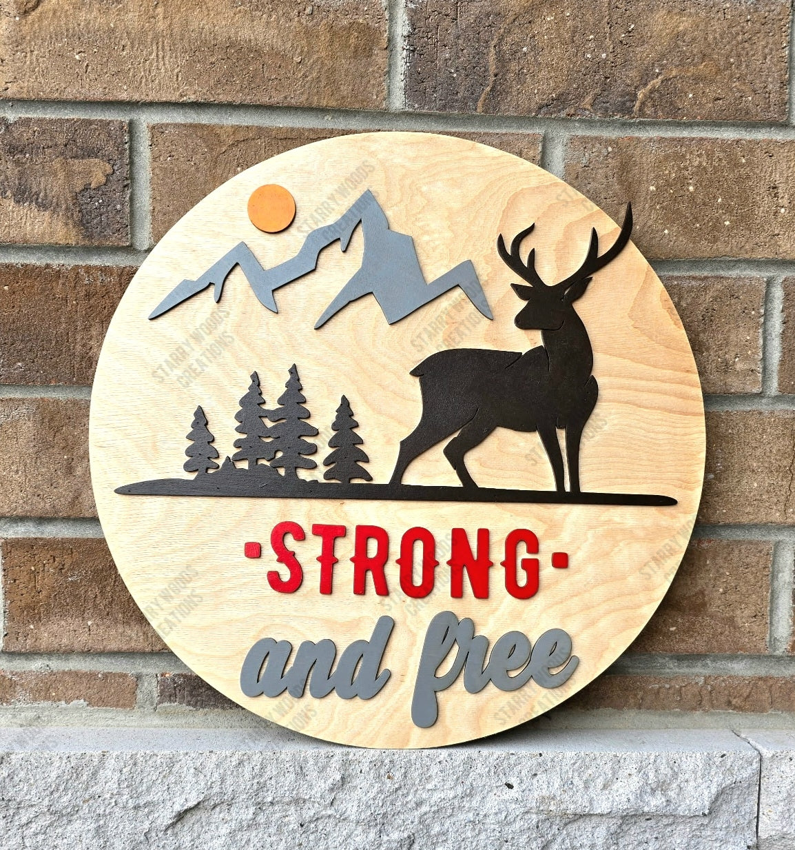 A round wooden door sign with a silhouette of a deer, mountains, and trees, featuring the phrases 'STRONG and free' in red and gray letters.