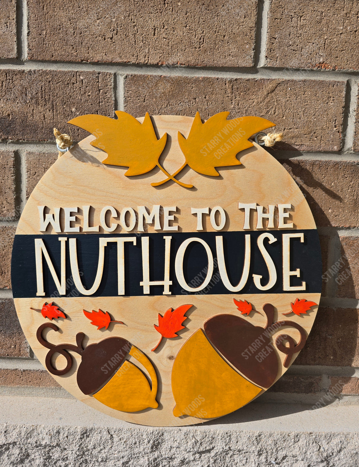 A circular wooden door sign with the text 'WELCOME TO THE NUTHOUSE' in capital letters, decorated with images of nuts and leaves in autumn colors.