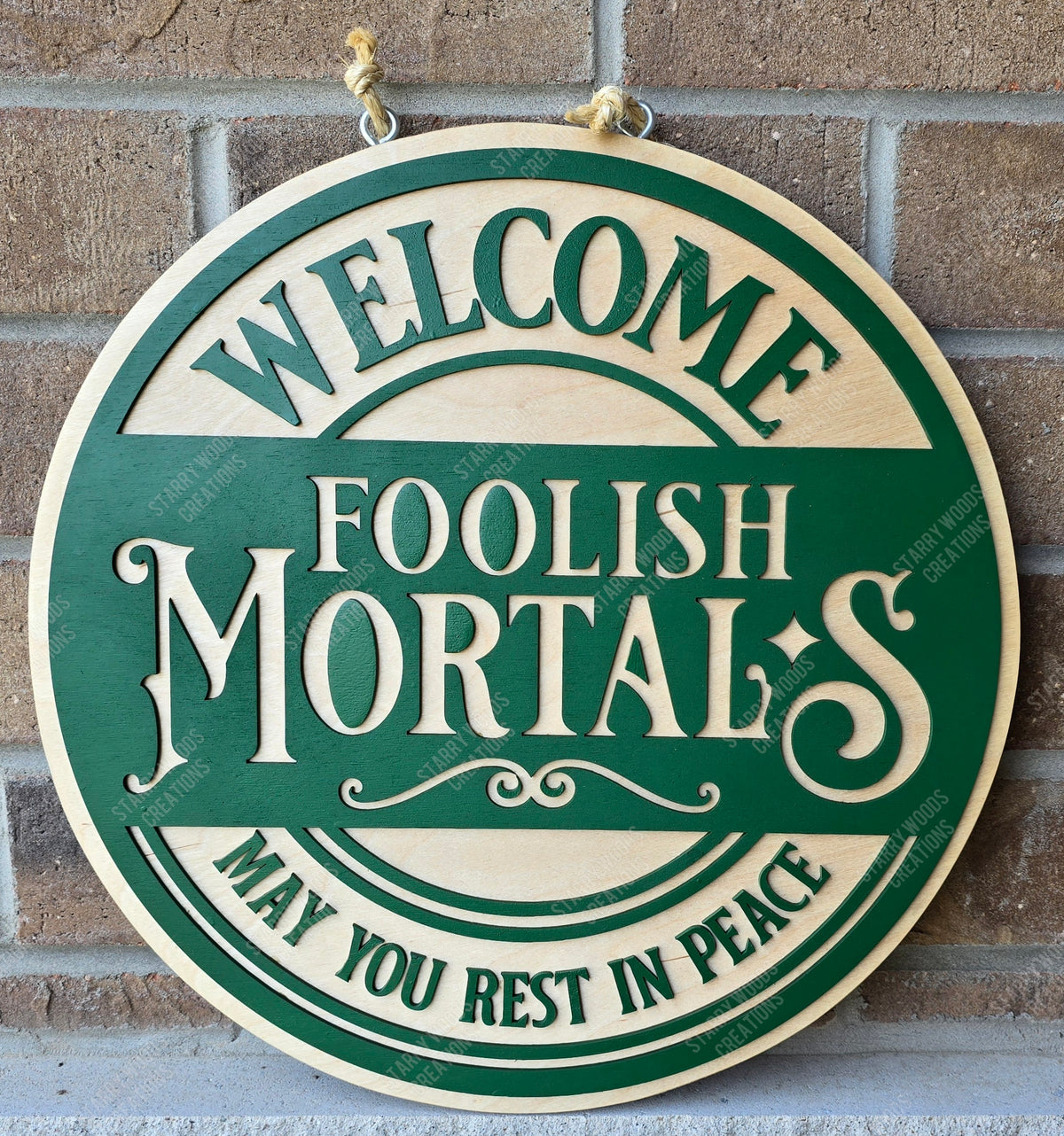 Round wooden sign with text 'Welcome Foolish Mortals May You Rest In Peace' on a brick wall.