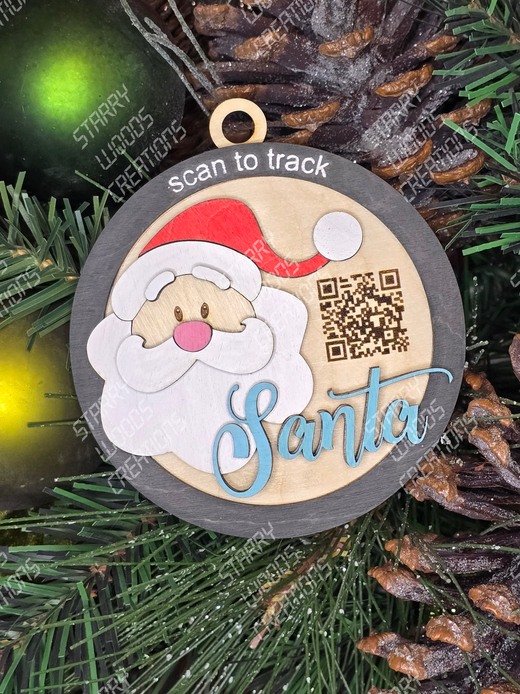 A round ornament with an illustration of Santa Claus and the text 'Santa', with a QR code above the text. The ornament is displayed against a background of green foliage.