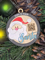A round ornament with an illustration of Santa Claus and the text 'Santa', with a QR code above the text. The ornament is displayed against a background of green foliage.