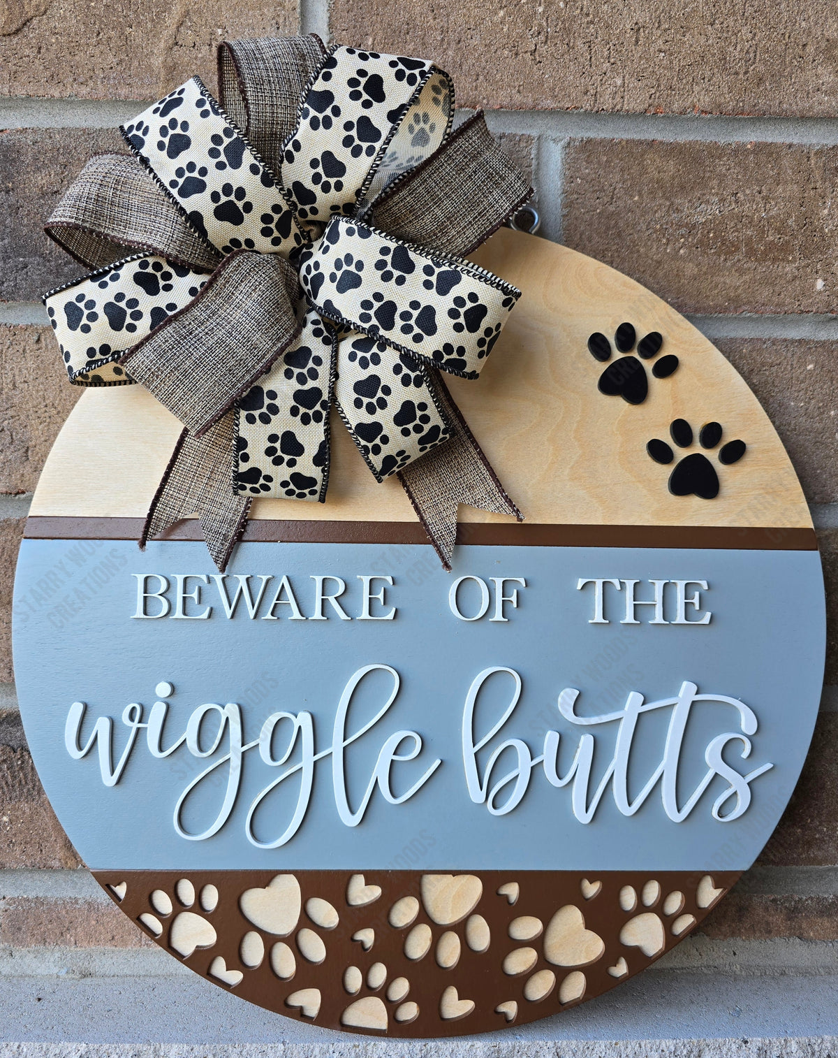 Decorative round sign with a paw print ribbon and 3D text reading 'Beware of the wiggle butts'. Layered wooden paw print designs and neutral colourings. Handmade in Quinte West, ready to hang and customizable.